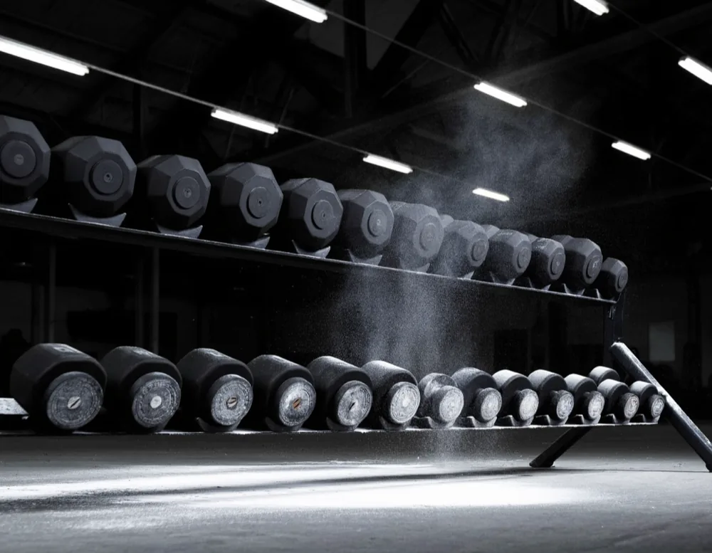 A two-tier dumbbell rack holding hexagonal rubber dumbbells, chalk dust in the light.