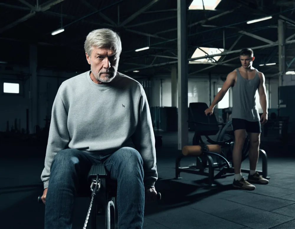 An older member on a cable row machine while a younger member spots a bench press behind him.